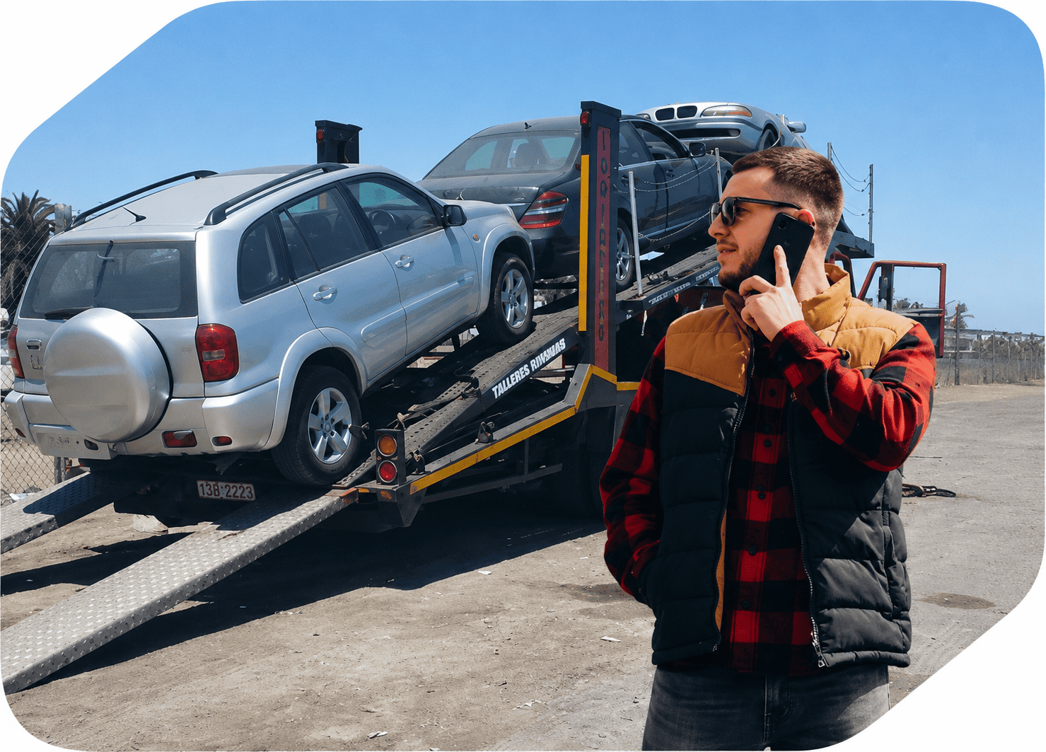 Car carrier truck transporting multiple vehicles while a logistics coordinator speaks on a mobile phone at a vehicle transport yard under a clear blue sky.